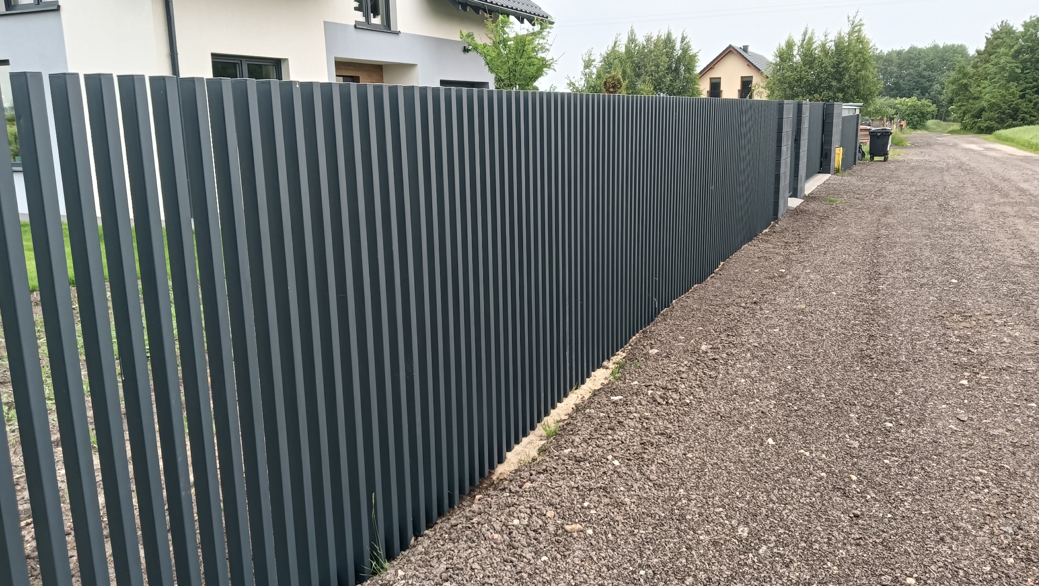 Nowoczesne, pionowe ogrodzenie metalowe w kolorze grafitowym, z widoczną fakturą. Tło stanowi dom i droga gruntowa. Ujęcie z perspektywy lekko z dołu.