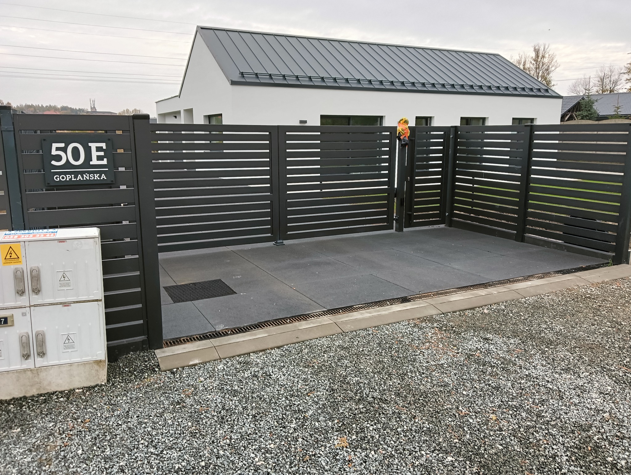 Nowoczesna, pozioma brama wjazdowa z metalowymi lamelami w kolorze antracytu, z numerem domu '50E Goplańska', na tle minimalistycznego, białego domu.