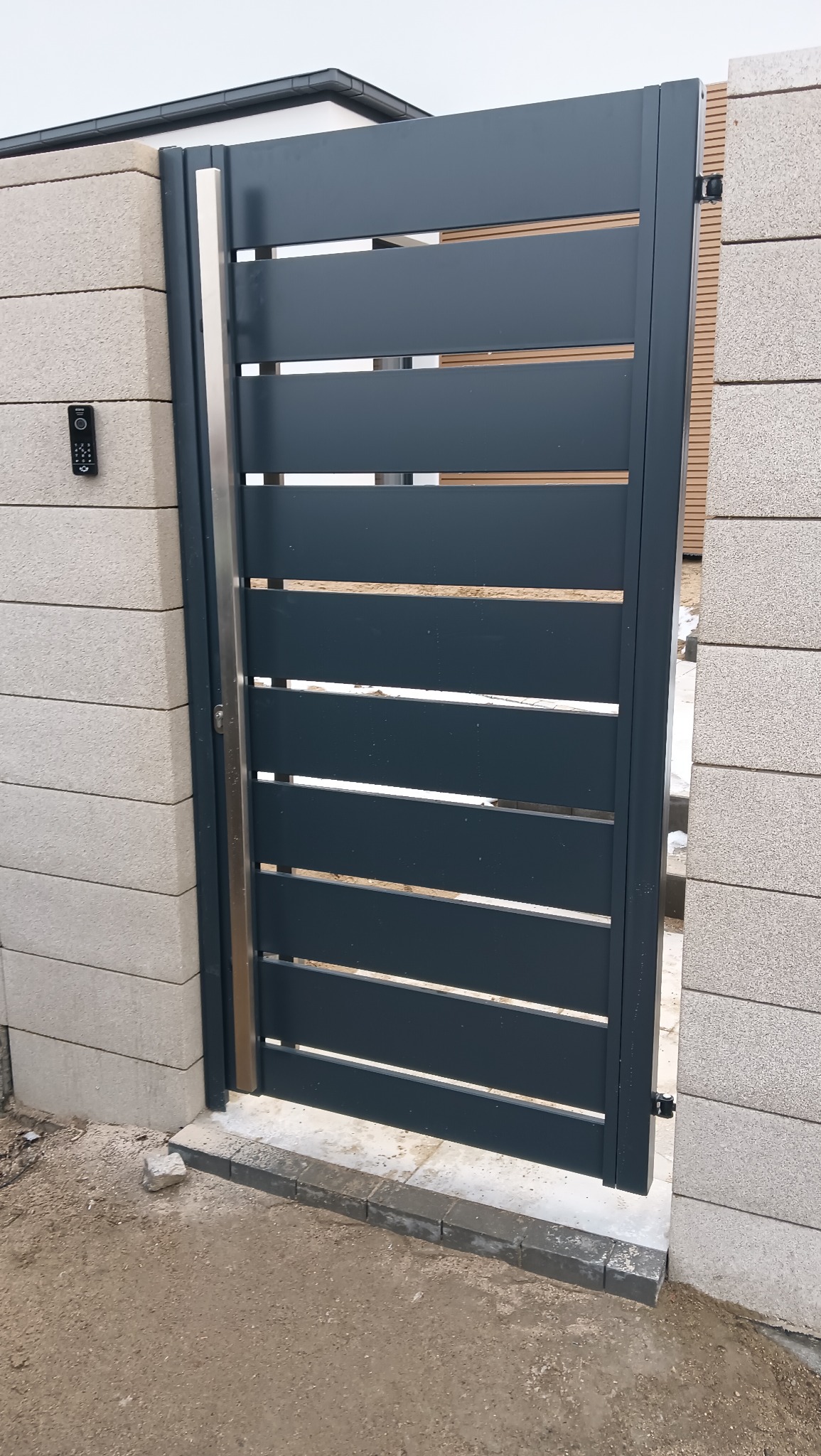 Nowoczesna brama wjazdowa z poziomych, grafitowych paneli, zamontowana na słupkach z szarego betonu architektonicznego. Widoczny domofon i stalowe wykończenia.