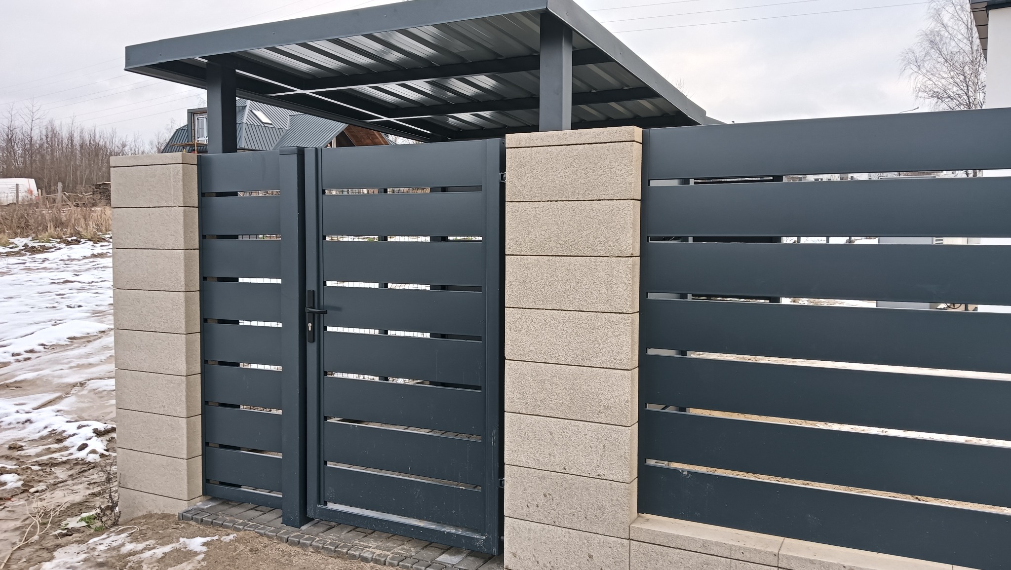 Nowoczesna, grafitowa brama wjazdowa z poziomymi szczeblami, osadzona na słupkach z jasnego kamienia. W tle widoczny zadaszony podjazd i fragment domu.