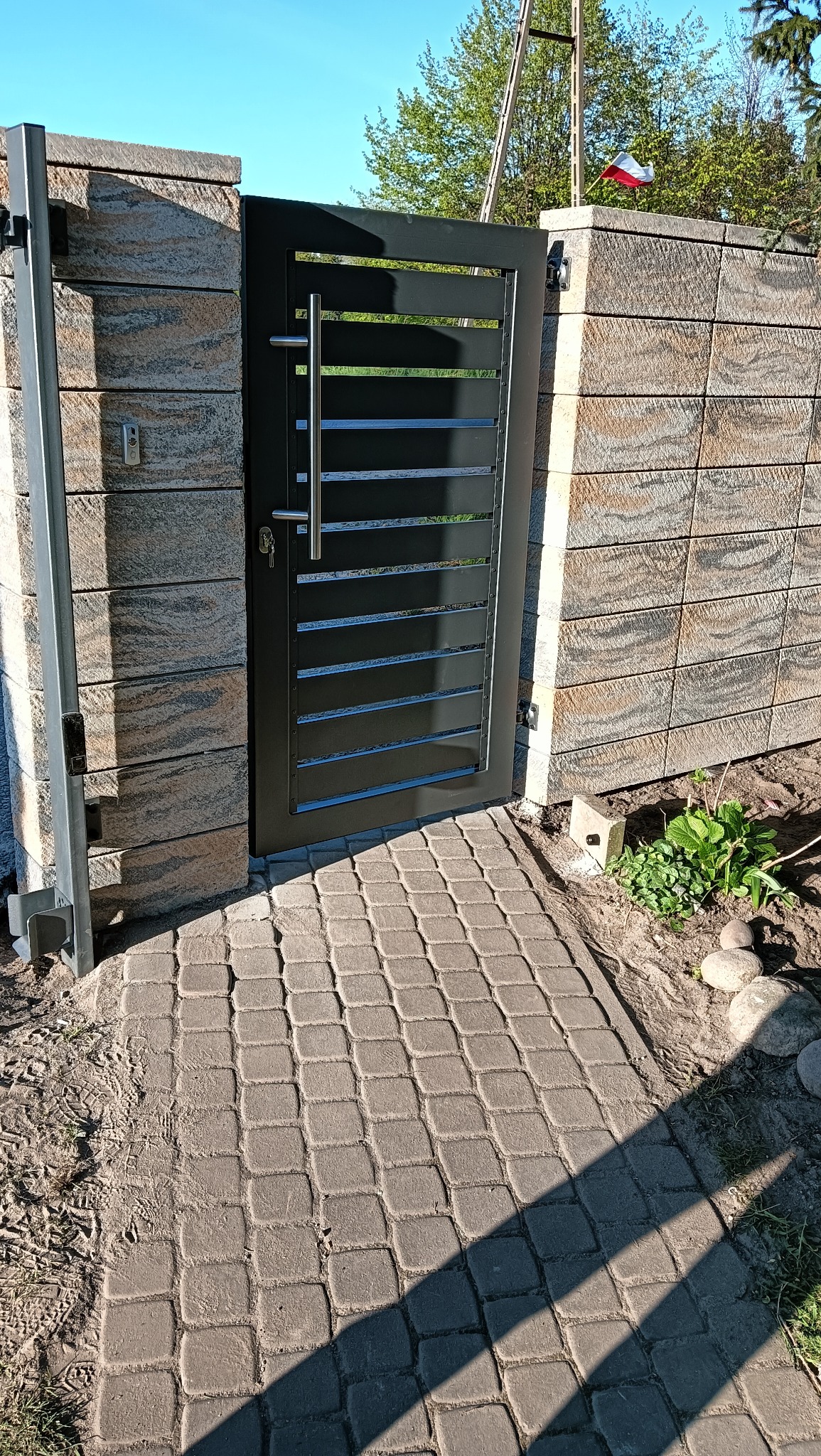 Nowoczesna, czarna furtka z poziomymi szczeblami i długim uchwytem, osadzona w murze z bloków imitujących kamień. Kostka brukowa prowadzi do wejścia.