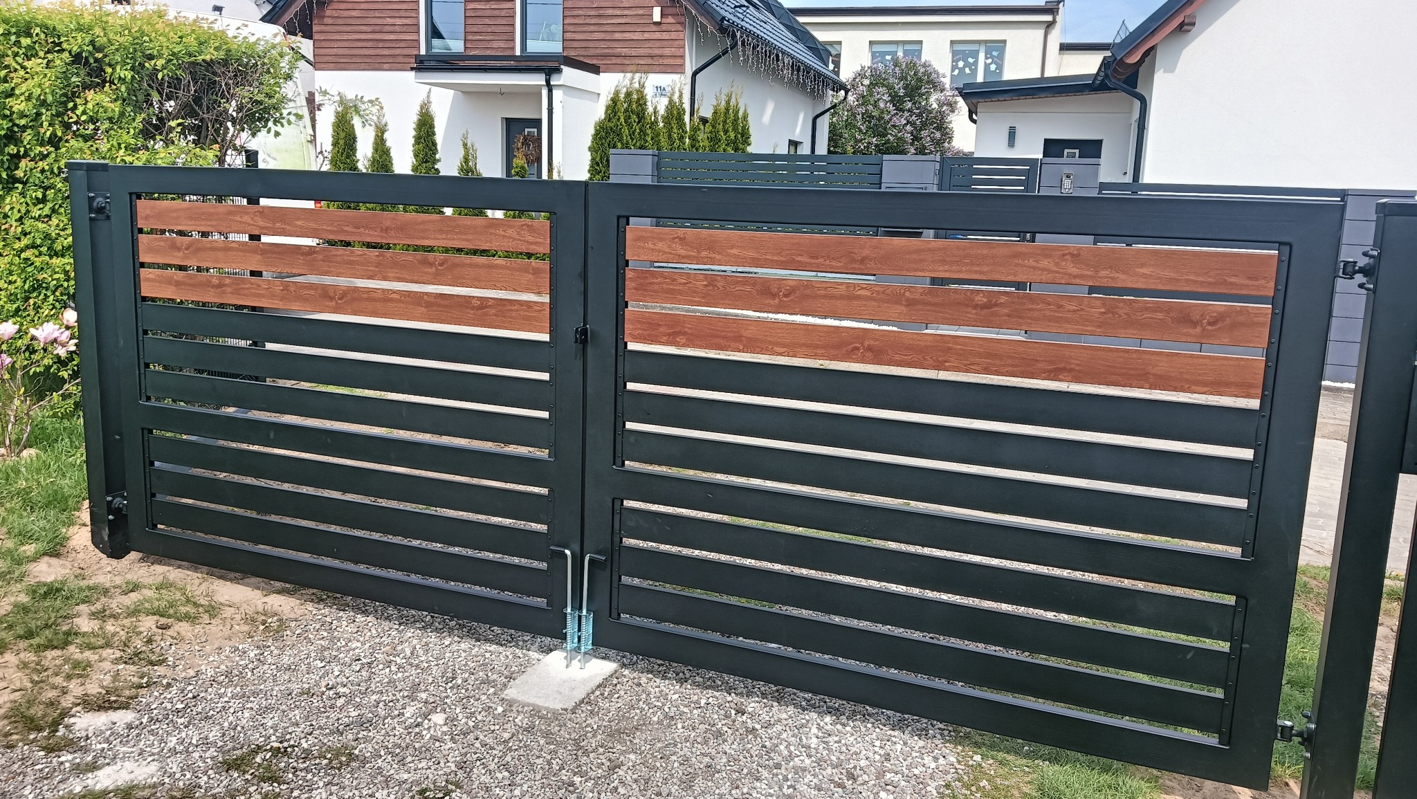 Nowoczesna brama wjazdowa z czarnych, poziomych profili stalowych i drewnopodobnych akcentów, na tle domów jednorodzinnych i żwirowego podjazdu.