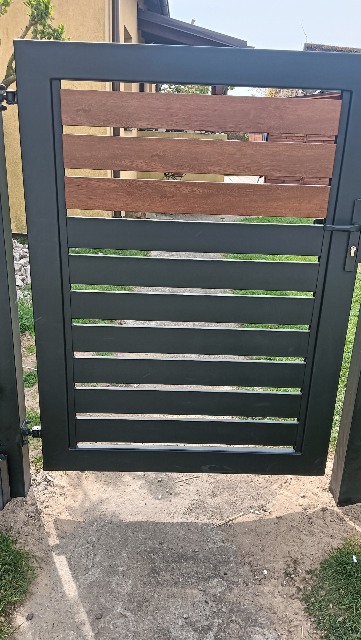 Nowoczesna brama wjazdowa z czarną, metalową ramą i poziomymi wypełnieniami z drewna kompozytowego. Widoczne zawiasy i zamek. Tło stanowi trawnik i fragment budynku.