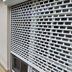 HIT PLUS - Biała, metalowa krata zabezpieczająca okno, częściowo opuszczona, z widocznymi szybami okiennymi w tle. Zewnętrzny montaż na tle beżowej elewacji.