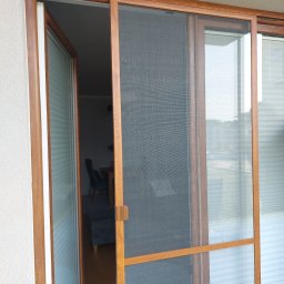 HIT PLUS - Przesuwana moskitiera w drewnianej ramie, zamontowana na balkonie. Widoczna faktura siatki i uchwyt. W tle wnętrze mieszkania z meblami.