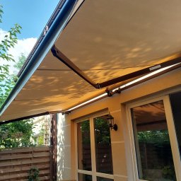 HIT PLUS - Beżowa markiza tarasowa rozłożona nad stołem ogrodowym, widok z dołu, błękitne niebo w tle. Oświetlenie boczne podkreśla strukturę materiału.