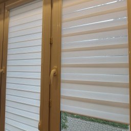 HIT PLUS - Beżowe okno z zamontowanymi, częściowo opuszczonymi białymi roletami dzień i noc. Widok na zewnątrz przez odsłoniętą część okna.