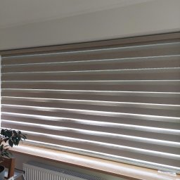 HIT PLUS - Beżowe rolety dzień noc z poziomymi pasami, zamontowane w oknie. Na parapecie stoi roślina doniczkowa, a pod nim widoczny jest biały grzejnik.