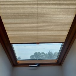 HIT PLUS - Beżowa, plisowana osłona okienna na oknie dachowym z drewnianą ramą, widok z dołu. Częściowo zasłonięte okno z widokiem na zewnątrz.