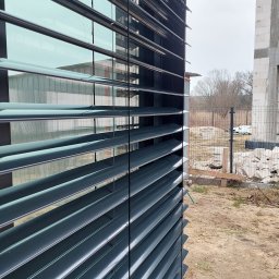 HIT PLUS - Zewnętrzne żaluzje fasadowe w kolorze grafitowym, zamontowane na budynku w trakcie budowy. Widoczne elementy konstrukcyjne i surowe otoczenie.