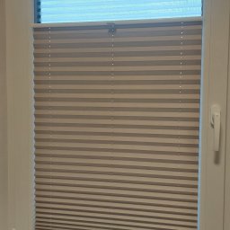 HIT PLUS - Beżowe plisy okienne zamontowane w oknie PCV, częściowo zasłonięte, z dekoracjami na parapecie: rośliny doniczkowe, perfumy i ozdobne słoneczniki.