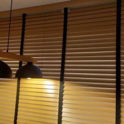 HIT PLUS - Drewniane żaluzje poziome w ciepłym odcieniu brązu z czarnymi taśmami, oświetlone przez wiszące lampy, tworzą przytulną atmosferę wnętrza.