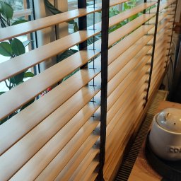 HIT PLUS - Drewniane żaluzje poziome z czarnym sznurkiem na oknie, częściowo zasłonięte, z widocznymi roślinami w tle i fragmentem blatu z ceramicznym pojemnikiem.