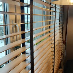 HIT PLUS - Drewniane żaluzje poziome z czarnymi taśmami na oknie, widoczne z bliska, z fragmentem lampy i dekoracyjnym pojemnikiem w kształcie misia w tle.