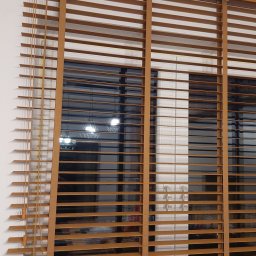 HIT PLUS - Drewniane żaluzje poziome w ciepłym odcieniu brązu, zamontowane w oknie. Widoczne sznurki sterujące i elementy mocujące. W tle rozmyte światła wnętrza.