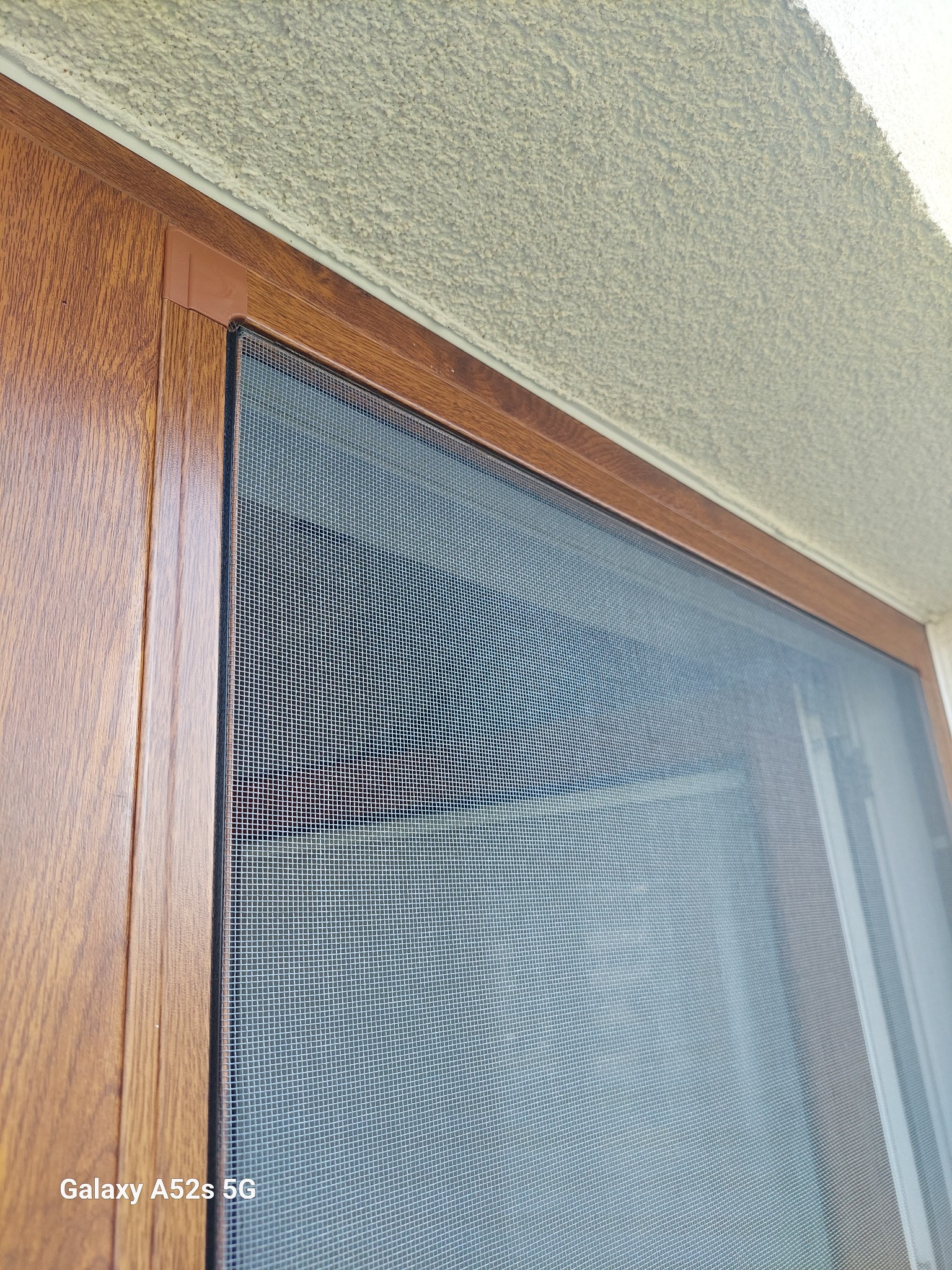 Zamknięta moskitiera w brązowej ramie, widoczna faktura siatki i drewnopodobne wykończenie ramy, z bliska, z delikatnym światłem padającym z góry.