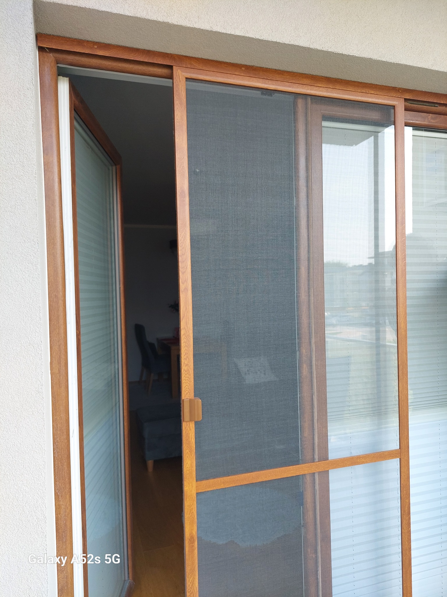 Przesuwana moskitiera w drewnianej ramie, zamontowana na balkonie. Widoczna faktura siatki i uchwyt. W tle wnętrze mieszkania z meblami.