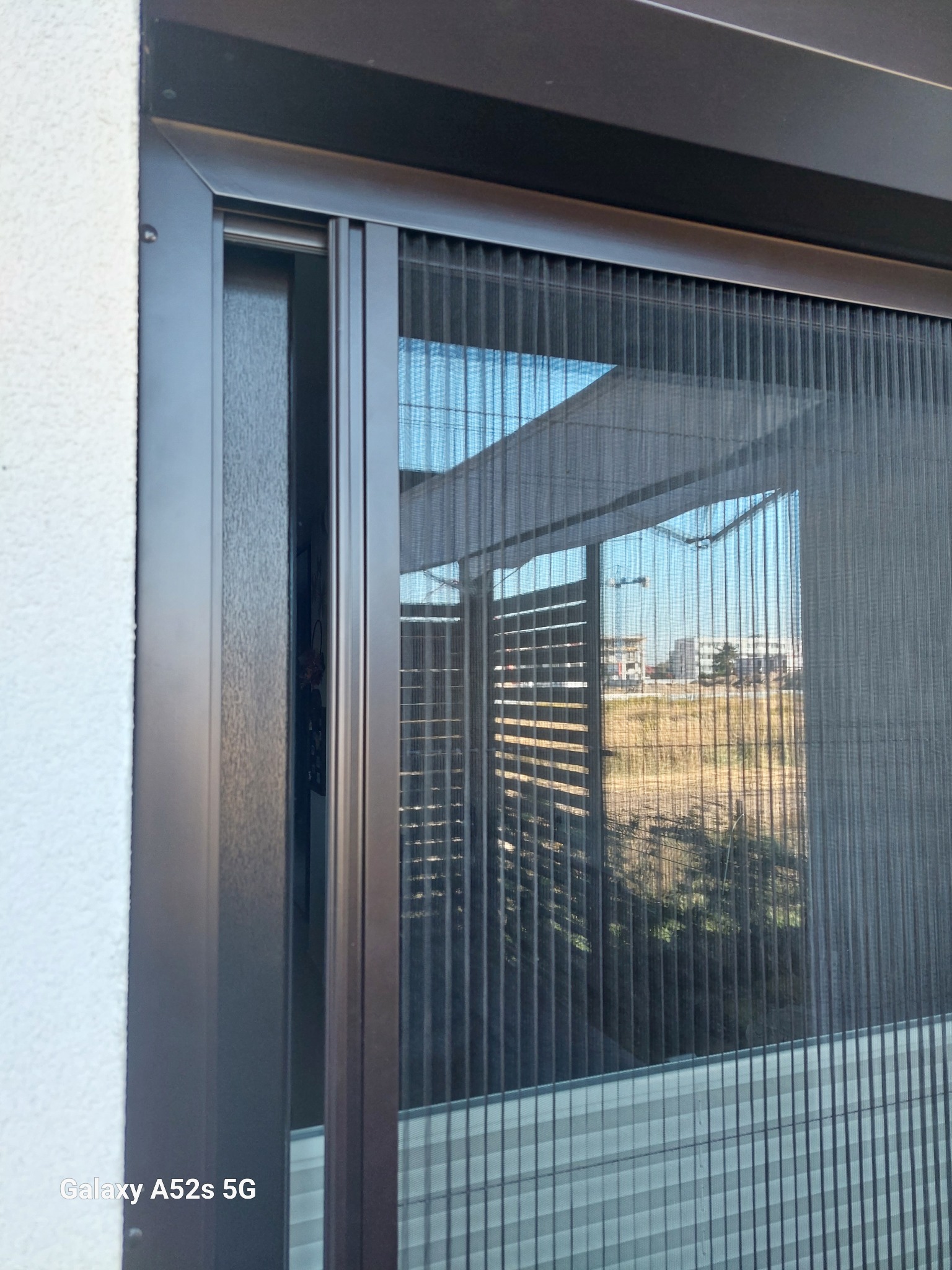 Zewnętrzna moskitiera plisowana w brązowej ramie, zamontowana na oknie. Widok na zewnątrz przez siatkę, zarys budynków w tle. Drobna faktura ściany po lewej.