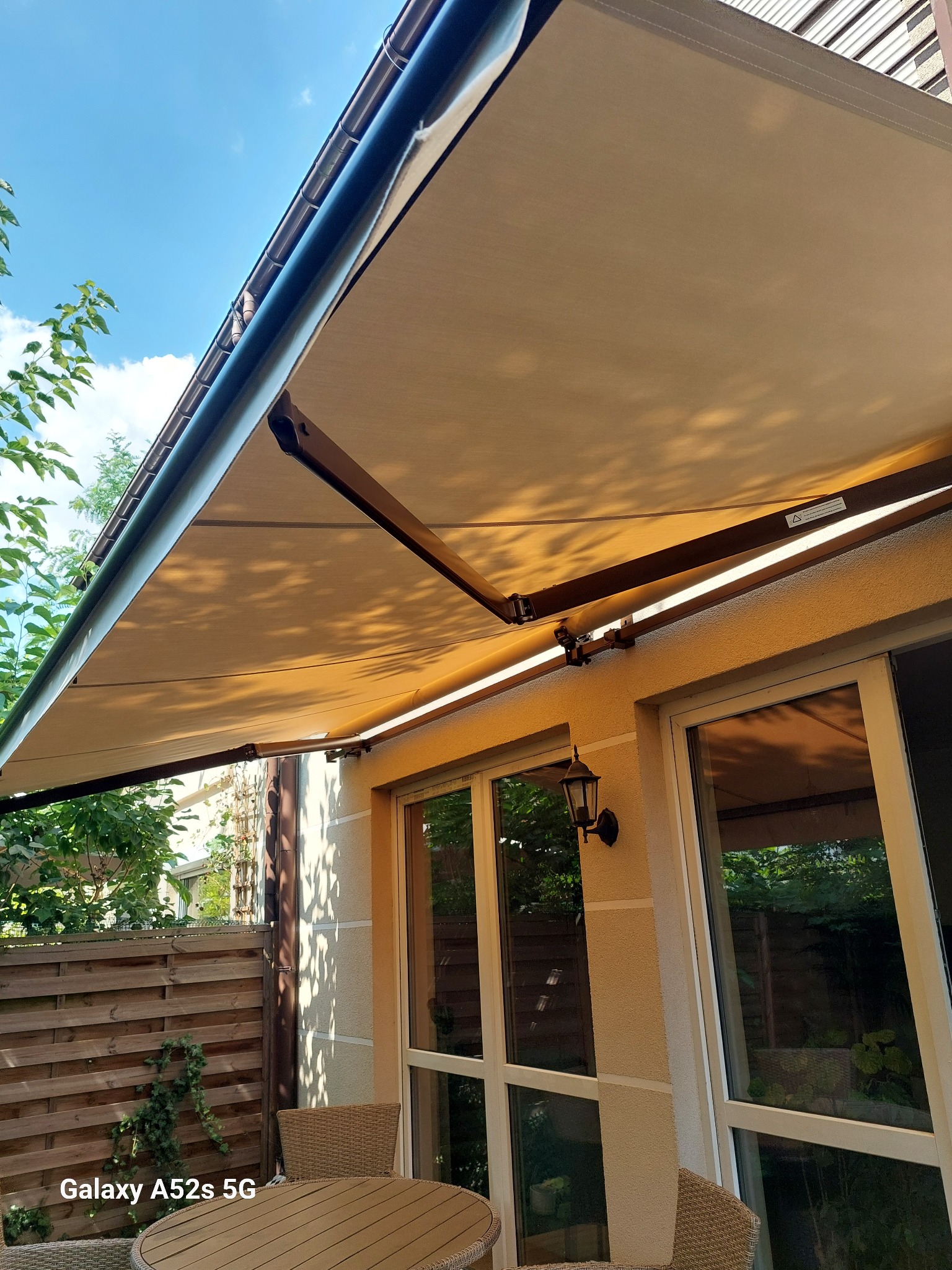 Beżowa markiza tarasowa rozłożona nad stołem ogrodowym, widok z dołu, błękitne niebo w tle. Oświetlenie boczne podkreśla strukturę materiału.