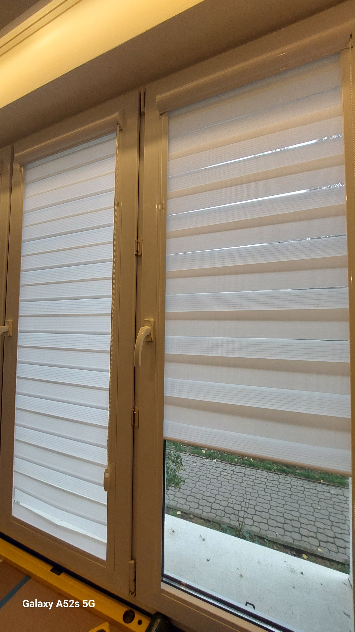 Beżowe okno z zamontowanymi, częściowo opuszczonymi białymi roletami dzień i noc. Widok na zewnątrz przez odsłoniętą część okna.