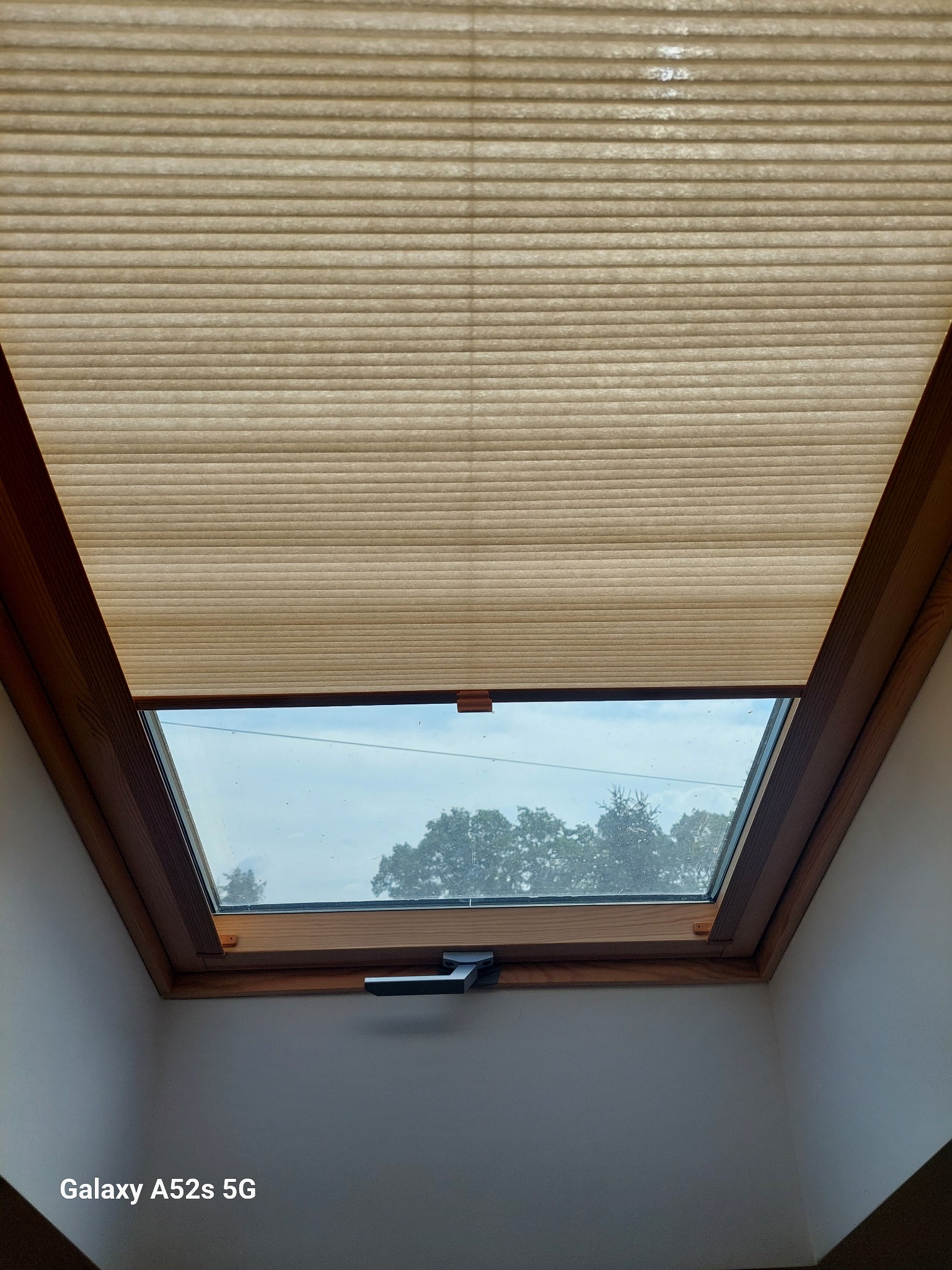 Beżowa, plisowana osłona okienna na oknie dachowym z drewnianą ramą, widok z dołu. Częściowo zasłonięte okno z widokiem na zewnątrz.