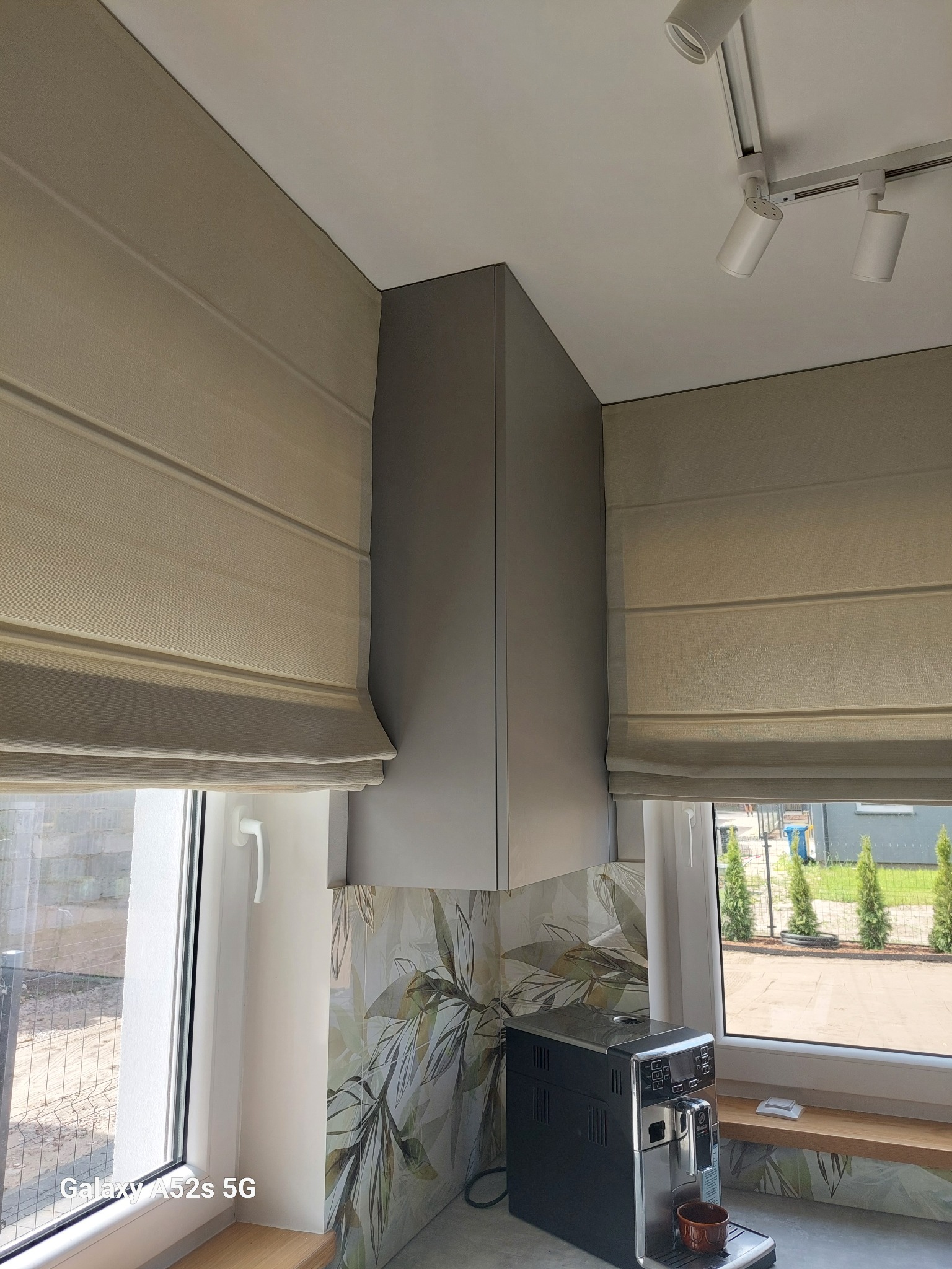 Beżowe rolety w oknach kuchni z wzorzystą glazurą i ekspresem do kawy. Widok na zewnątrz z zielenią. Oświetlenie sufitowe punktowe.