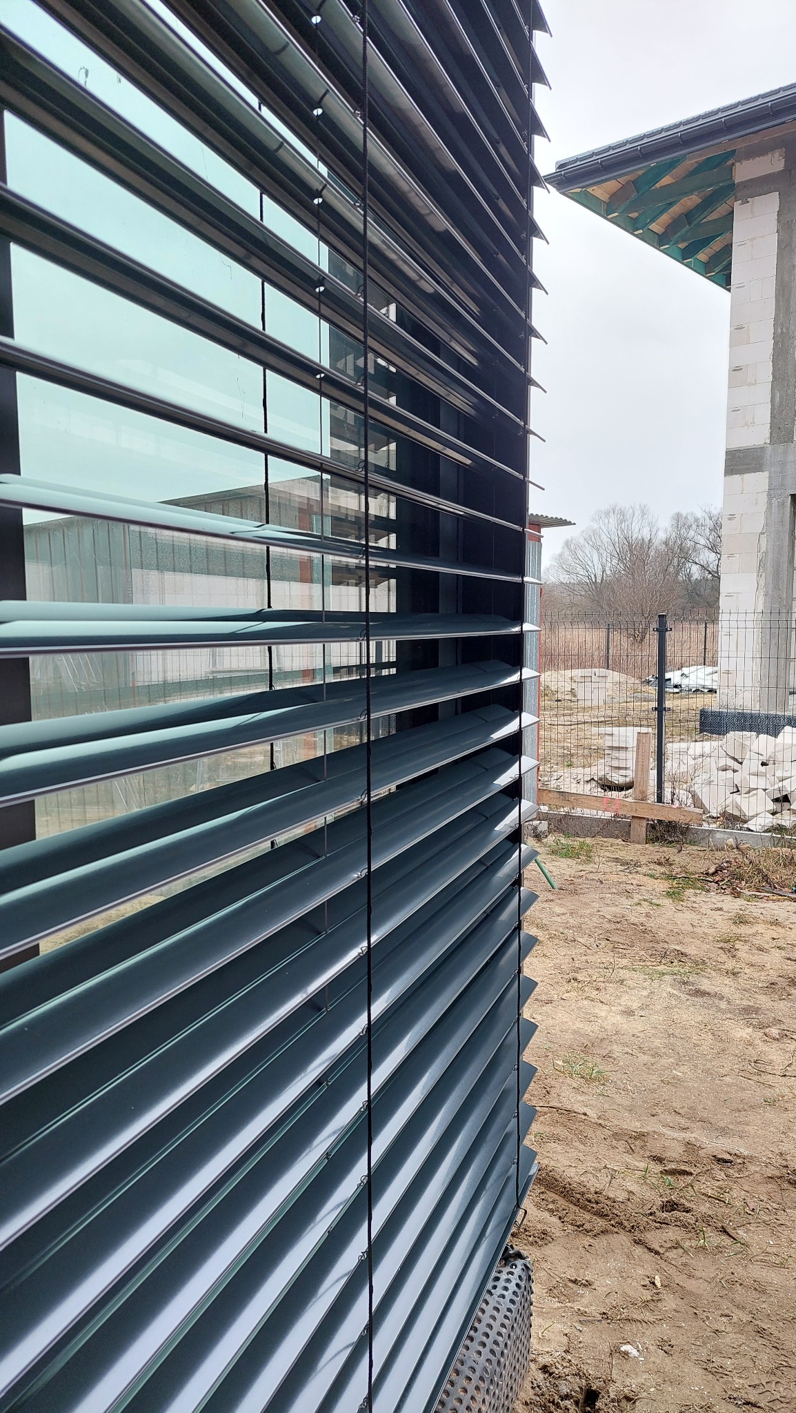 Zewnętrzne żaluzje fasadowe w kolorze grafitowym, zamontowane na budynku w trakcie budowy. Widoczne elementy konstrukcyjne i surowe otoczenie.