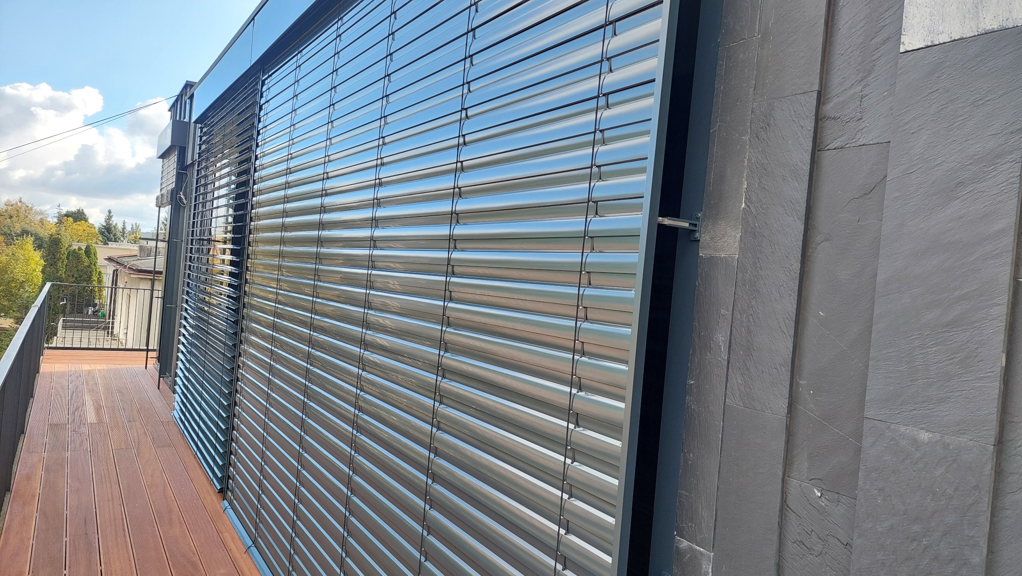 Metalowe żaluzje zewnętrzne zamontowane na balkonie z drewnianą podłogą, widok z boku, z elementami elewacji z kamienia łupkowego w tle.