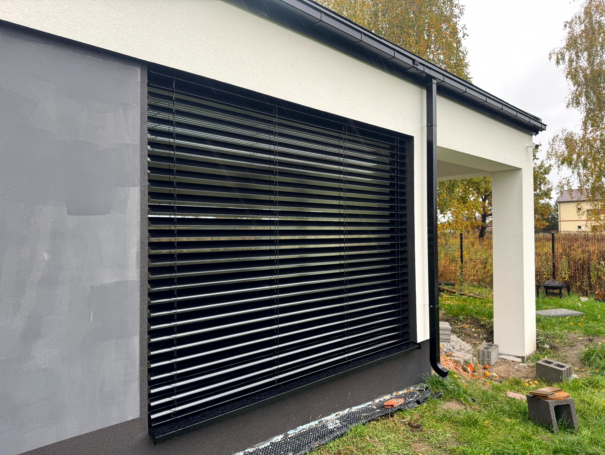Czarne poziome lamele okienne na tle szarej i białej elewacji budynku. Widoczne elementy drenażu przy gruncie i fragment ogrodu.
