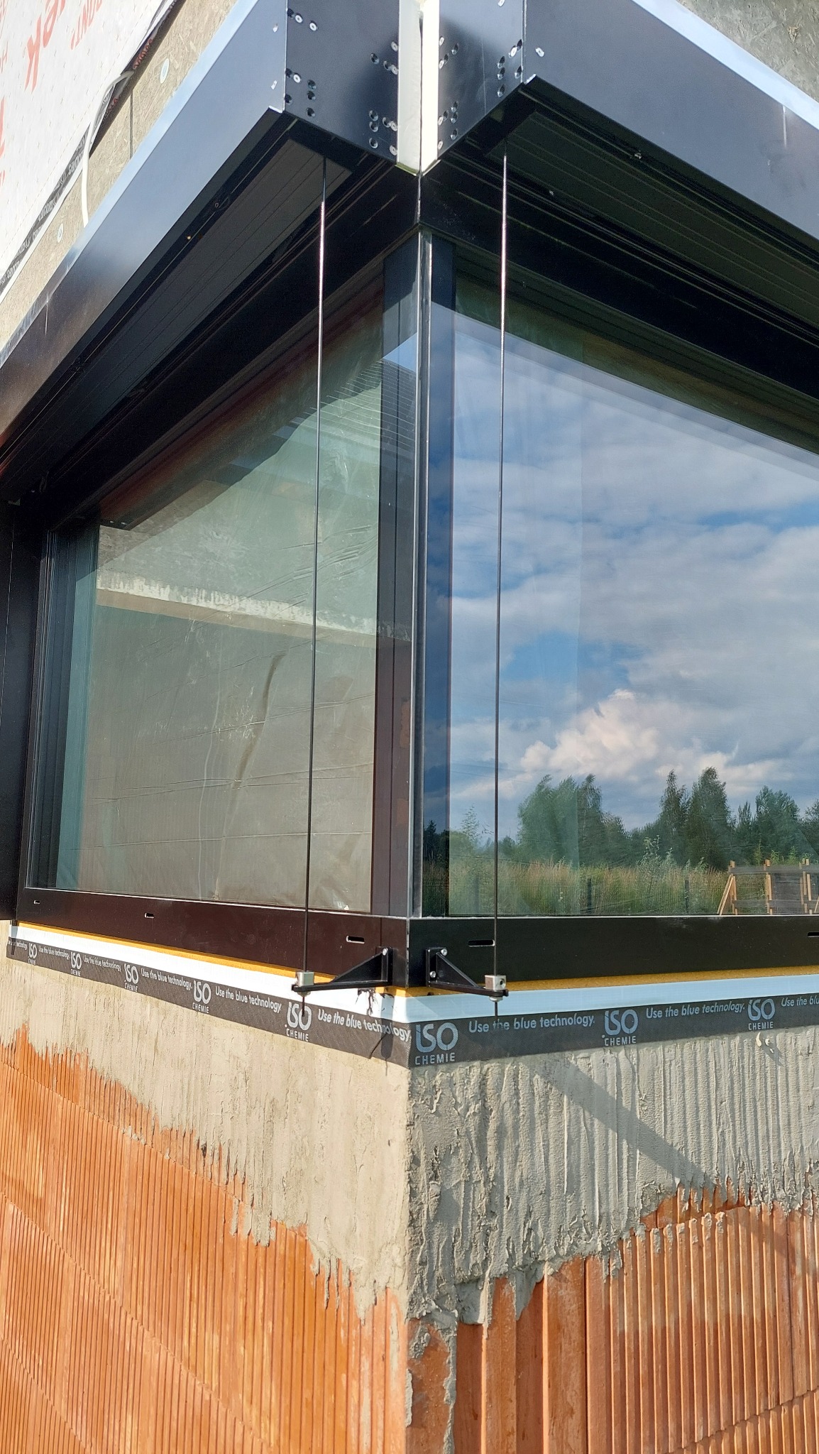 Nowoczesne, narożne okno z czarną ramą i widokiem na zewnątrz, zamontowane na surowej, ceglanej ścianie z widoczną izolacją. Detal architektoniczny.