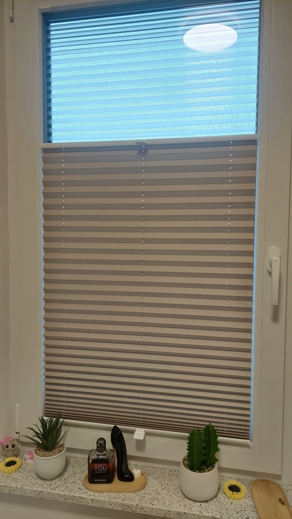 Beżowe plisy okienne zamontowane w oknie PCV, częściowo zasłonięte, z dekoracjami na parapecie: rośliny doniczkowe, perfumy i ozdobne słoneczniki.