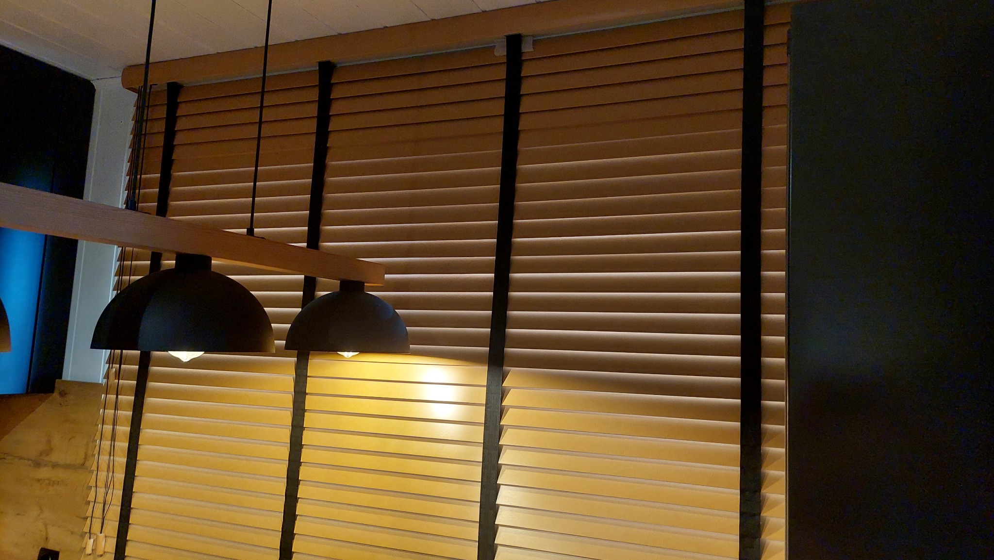 Drewniane żaluzje poziome w ciepłym odcieniu brązu z czarnymi taśmami, oświetlone przez wiszące lampy, tworzą przytulną atmosferę wnętrza.