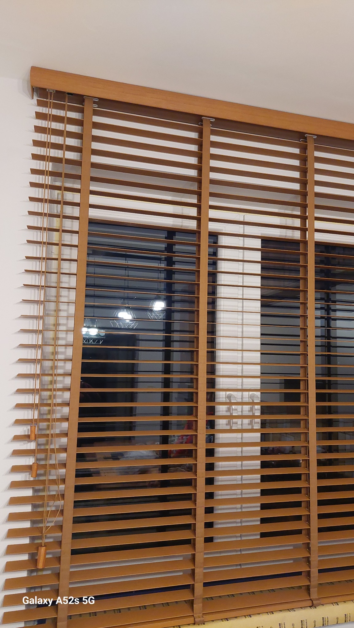 Drewniane żaluzje poziome w ciepłym odcieniu brązu, zamontowane w oknie. Widoczne sznurki sterujące i elementy mocujące. W tle rozmyte światła wnętrza.