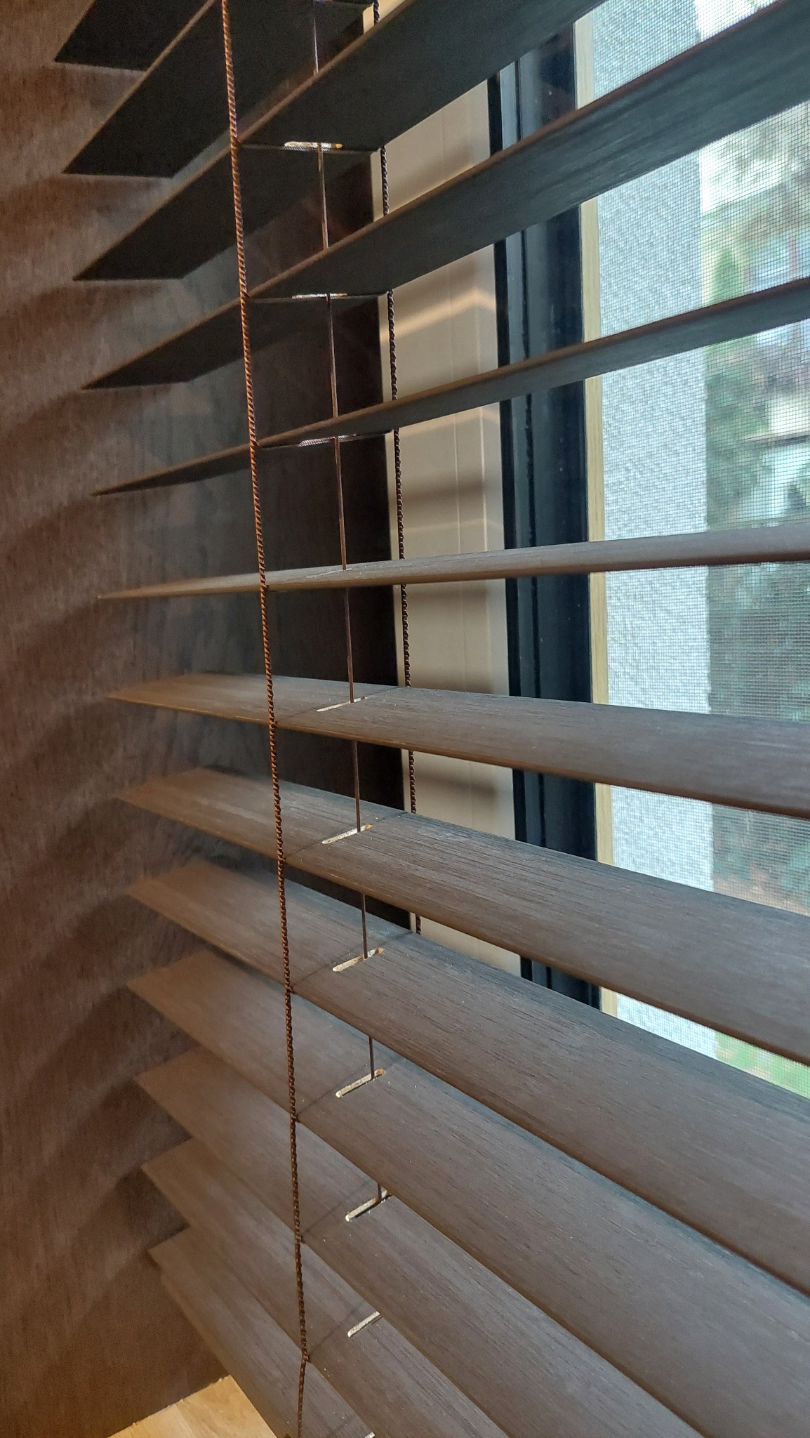 Drewniane żaluzje poziome w odcieniach brązu, częściowo zasłonięte, z widoczną strukturą drewna i sznurkami regulacyjnymi, na tle okna z moskitierą.