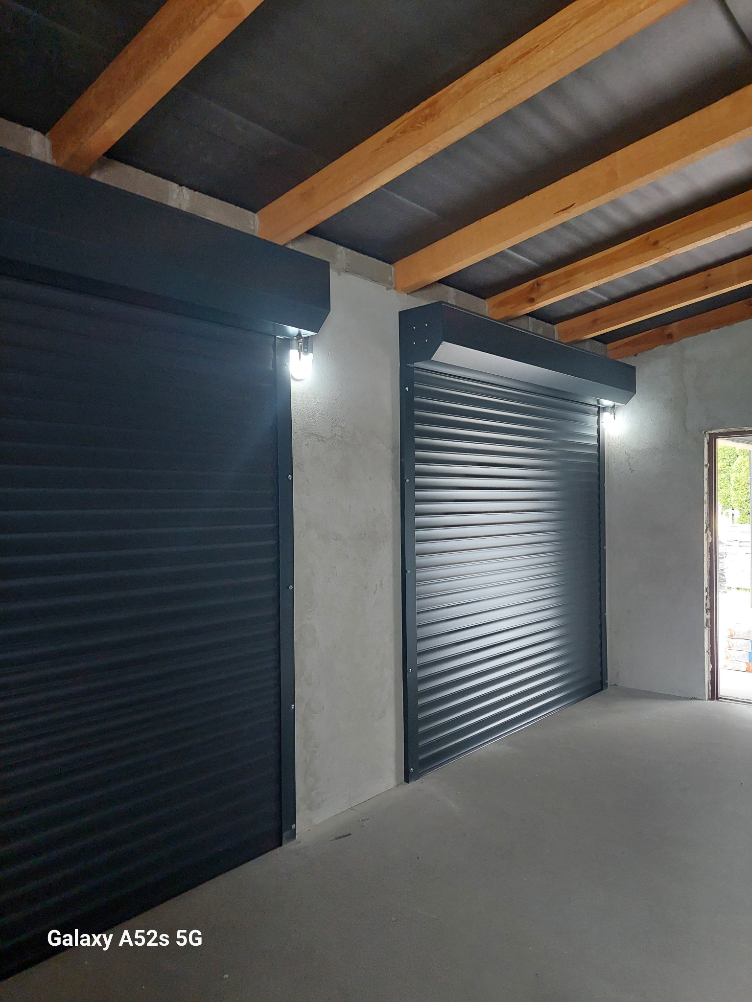 Dwie szare, metalowe osłony okienne w surowym wnętrzu z betonową podłogą i drewnianymi belkami stropowymi. Jedna osłona jest zamknięta, druga częściowo otwarta.