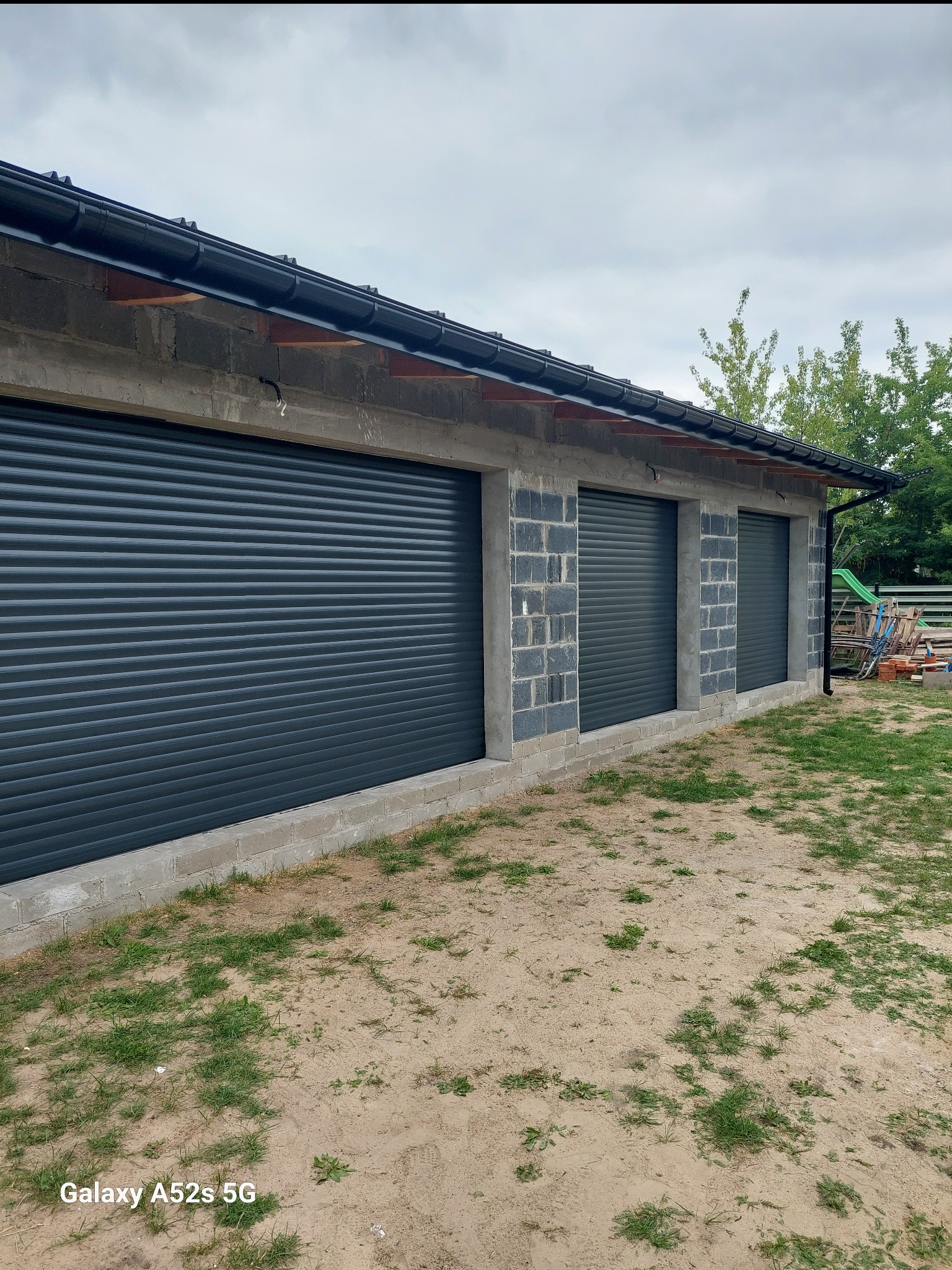 Szare, metalowe osłony okienne w surowym budynku z bloków betonowych. Widok z zewnątrz, na tle zieleni i pochmurnego nieba. Trzy okna z osłonami.