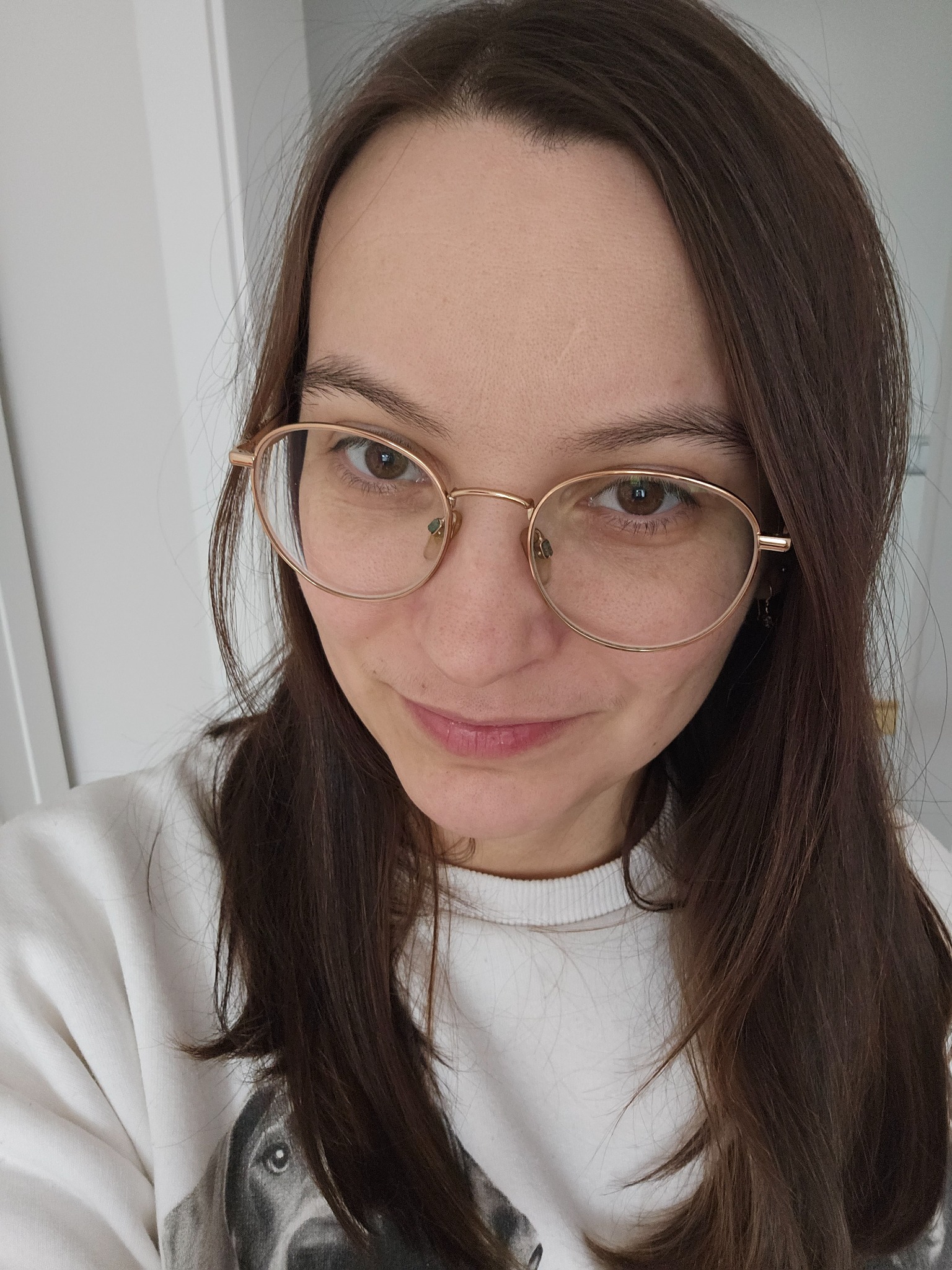 Portret kobiety w okularach o owalnych oprawkach, uśmiechającej się do kamery, w białej bluzie z nadrukiem psa, na tle białej ściany i drzwi.