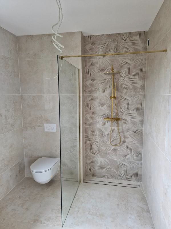 Wykończona łazienka z kabiną typu walk-in, złotą armaturą i toaletą podwieszaną. Płytki w odcieniach beżu i szarości z motywem liści. Widoczne przewody elektryczne.