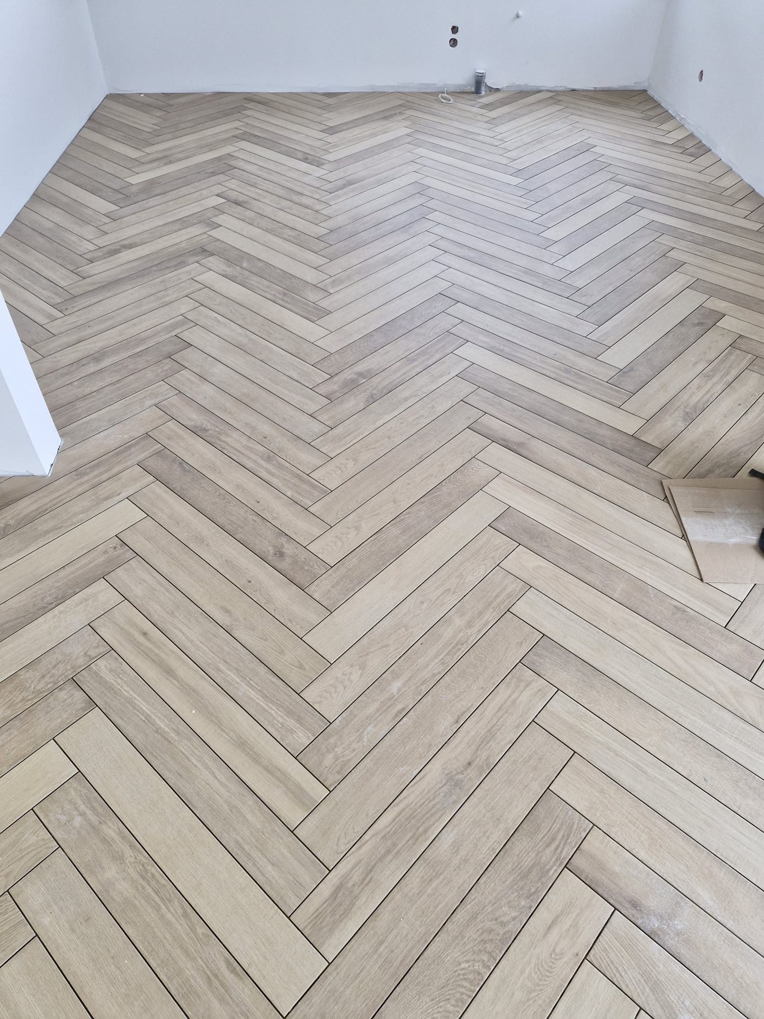Ułożona podłoga z płytek imitujących drewno w jodełkę, jasne fugi, widok z góry, fragment ściany w rogu. Częściowo widoczne płytki przed ułożeniem.