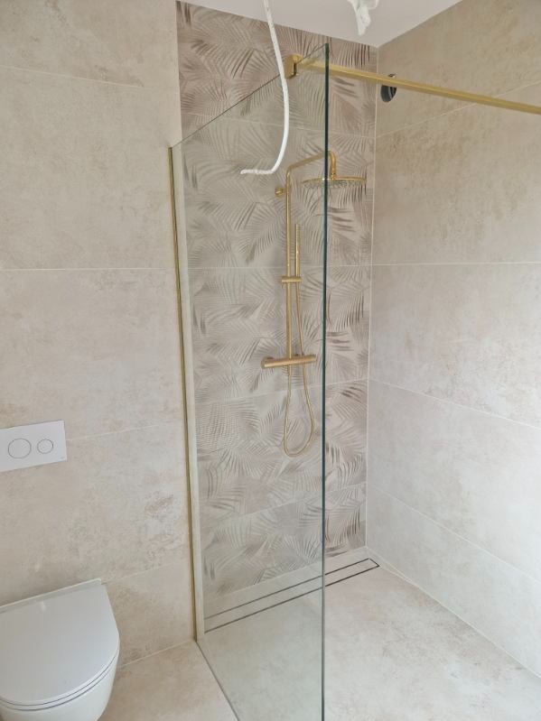 Beżowa łazienka z kabiną typu walk-in, złotą armaturą i płytkami w liście palmy. Widoczny fragment toalety i odpływ liniowy.