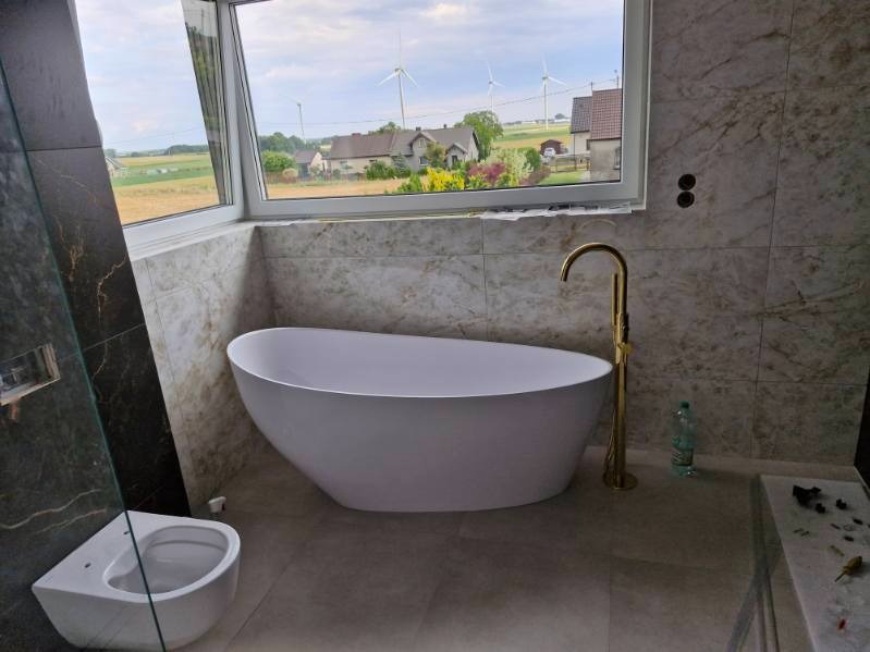 Nowoczesna łazienka z wolnostojącą białą wanną i złotą armaturą na tle okna z widokiem na krajobraz z turbinami wiatrowymi. Marmurowe płytki na ścianach.