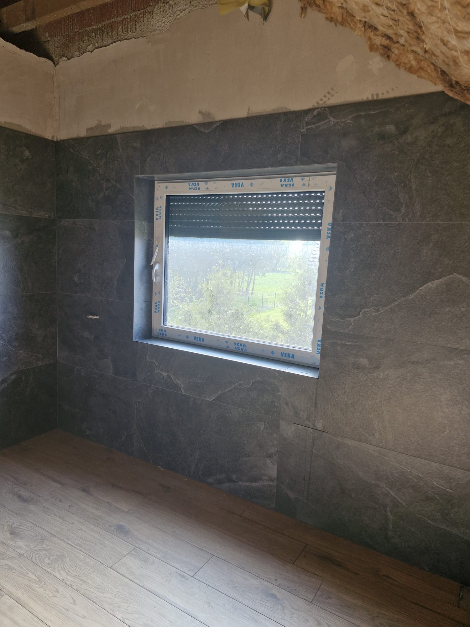 Wykończona łazienka z szarymi płytkami imitującymi kamień, drewnopodobną podłogą i oknem z roletą. Widoczne niedokończone wykończenie sufitu.