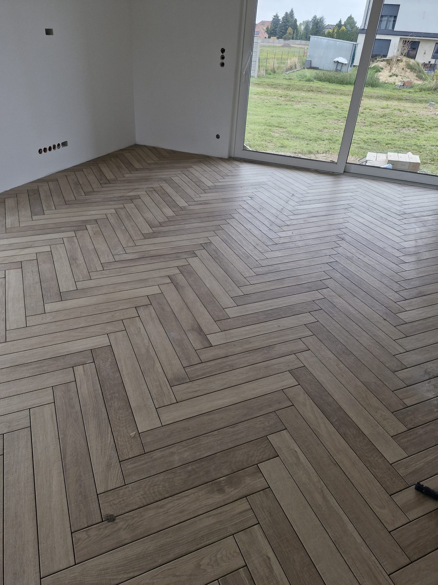 Ukończona podłoga z płytek imitujących drewno, ułożonych we wzór jodełki francuskiej. Widoczne gniazdka elektryczne na ścianie i duże okno z widokiem na zewnątrz.