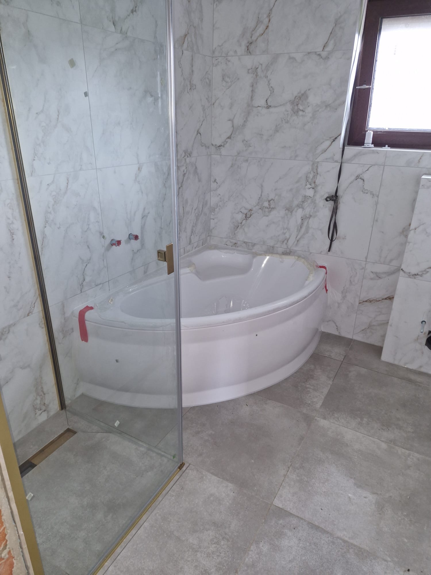 Nowoczesna łazienka z narożną wanną i kabiną prysznicową typu walk-in. Marmurowe płytki ścienne i szare płytki podłogowe. Widoczne elementy instalacji.