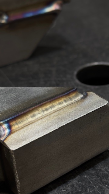 Zbliżenie na spaw na metalowym elemencie, widoczne tęczowe przebarwienia. Precyzyjne łączenie metali, detal spoiny na szarym tle warsztatowym.