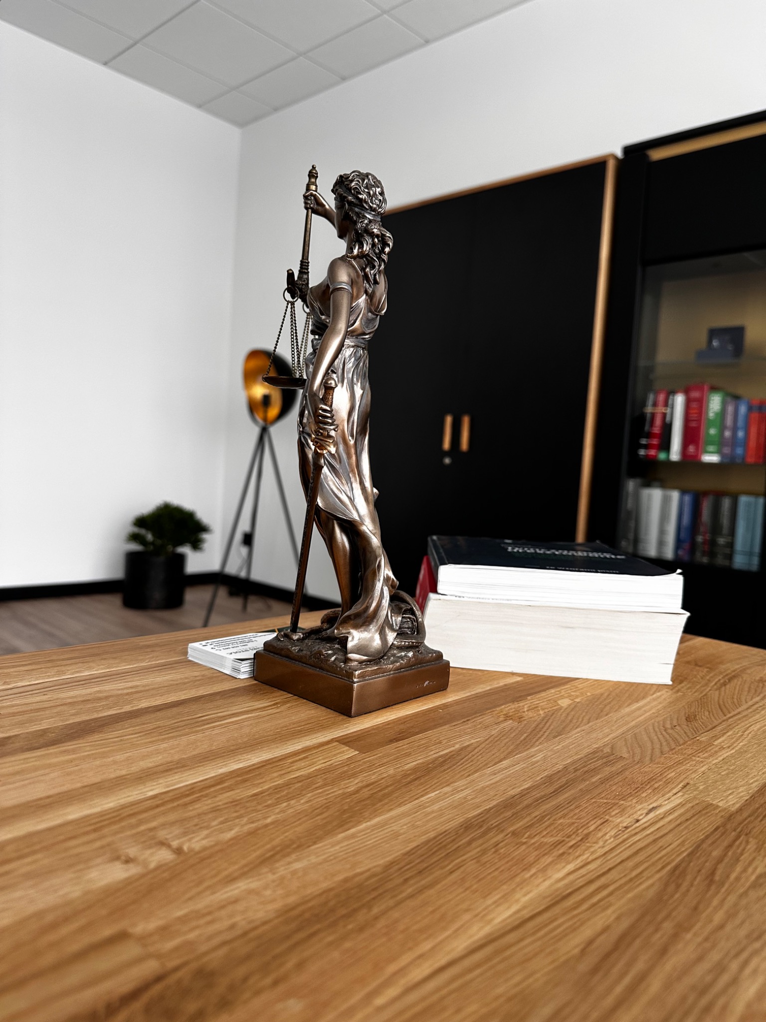 Statuetka Temidy na drewnianym biurku w kancelarii, obok wizytówki i książki. W tle regał z książkami i lampa na trójnogu.