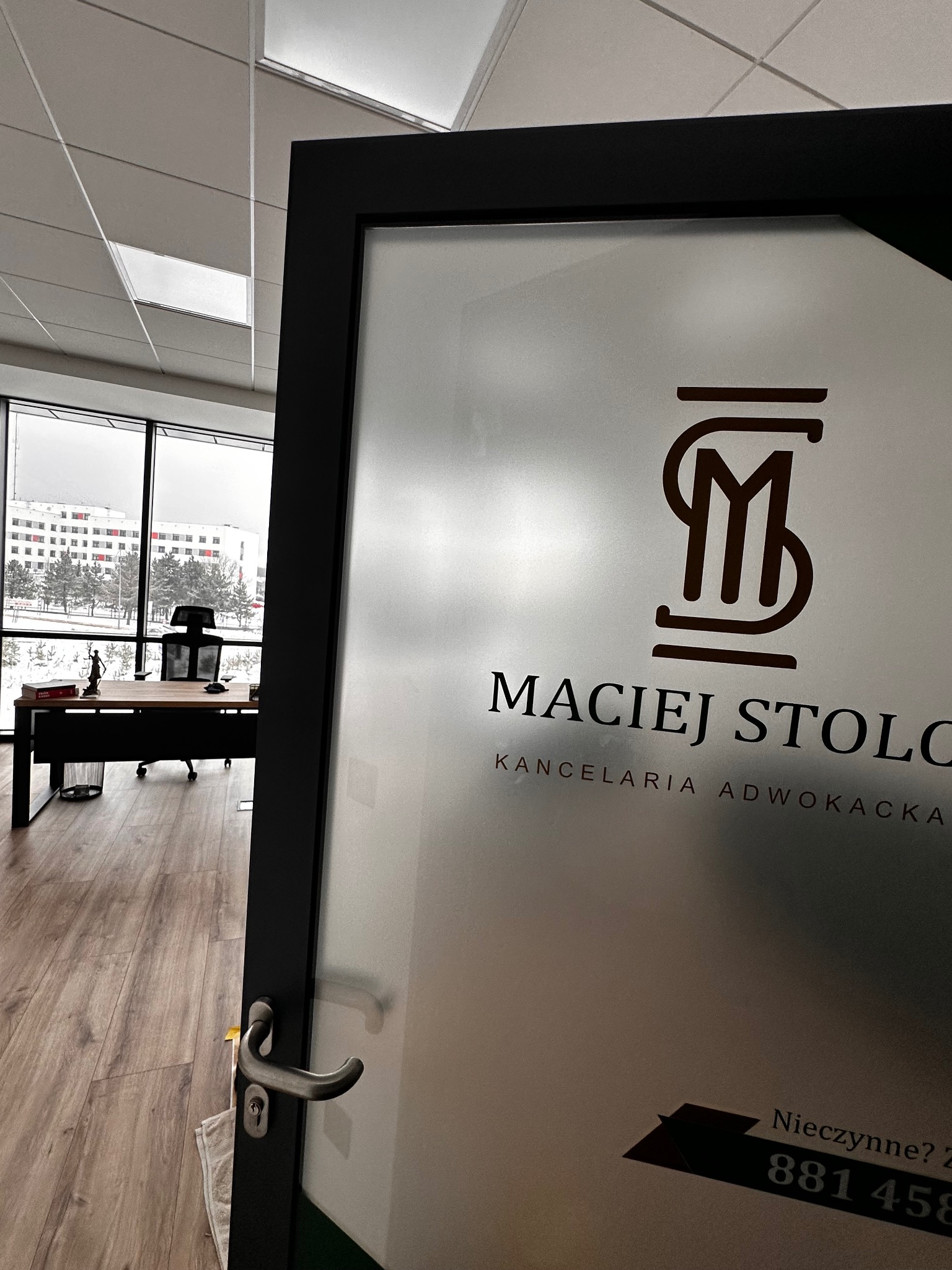 Szklane drzwi do kancelarii adwokackiej Maciej Stolc z logo i numerem telefonu, widok z wnętrza biura na biurko i okno z widokiem na zimowy krajobraz.