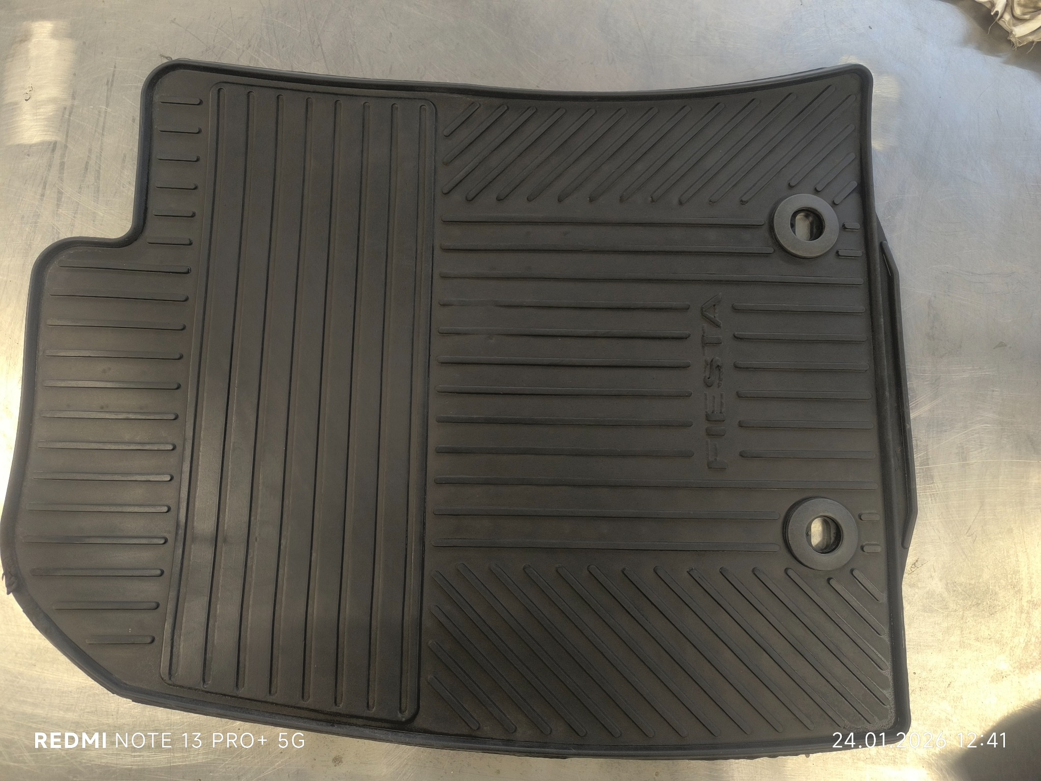 Czarna, gumowa wycieraczka samochodowa Ford Fiesta z wyraźnym wzorem i dwoma otworami montażowymi, leżąca na metalowej powierzchni.