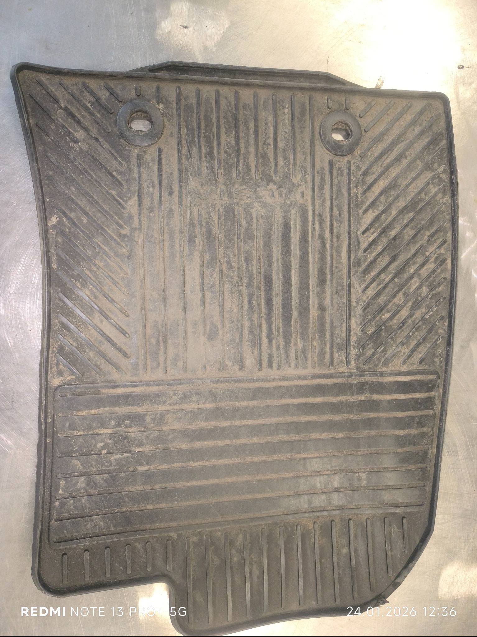 Brudny, gumowy dywanik samochodowy marki Volvo, z widocznymi otworami montażowymi i wzorem antypoślizgowym, leżący na metalowej powierzchni. Widoczne ślady użytkowania.