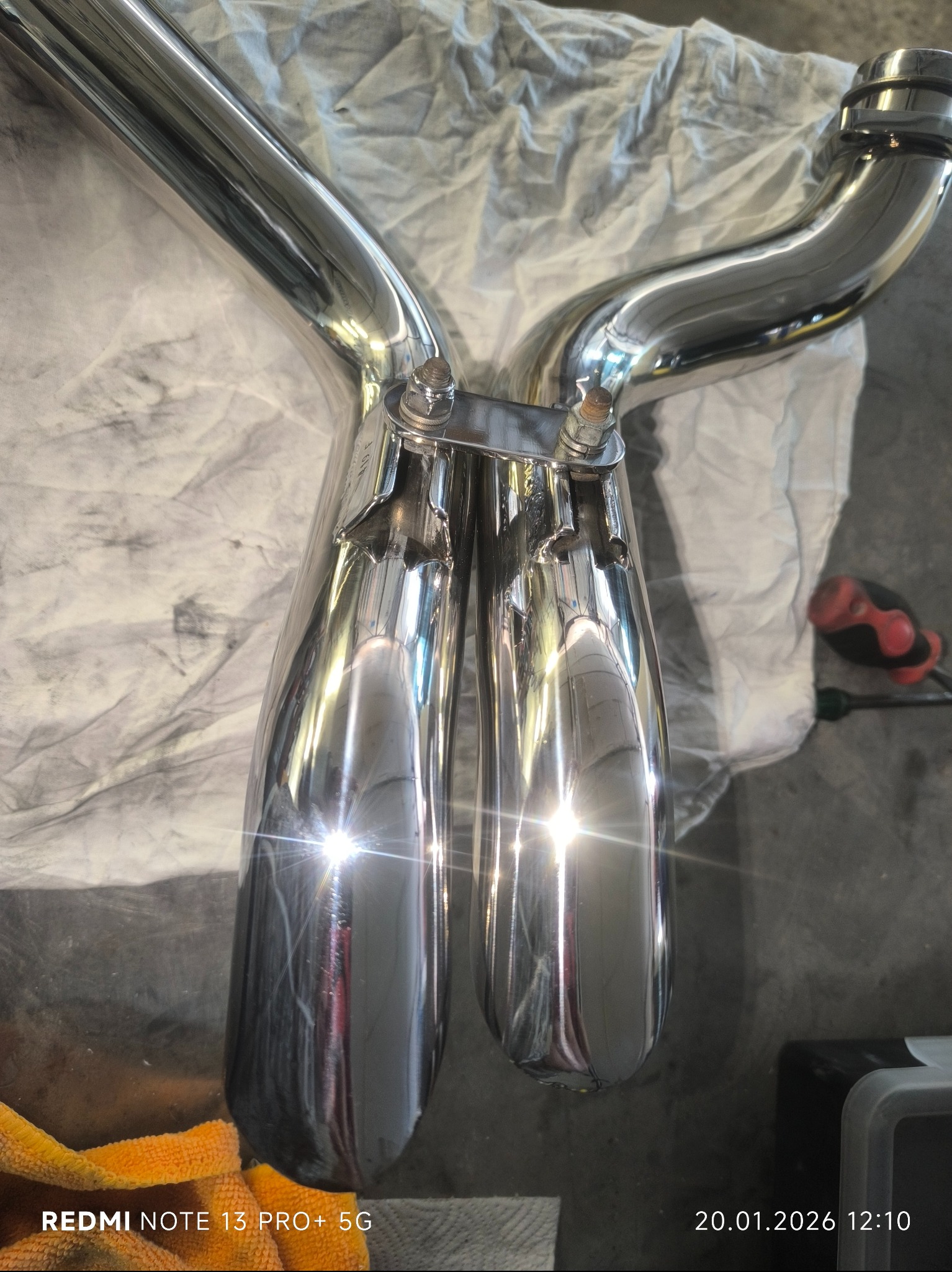 Błyszczący, chromowany układ wydechowy motocykla na tle szarej tkaniny, z widocznym kluczem i ściereczką. Detal wykonania i odbicia światła podkreślają jakość.